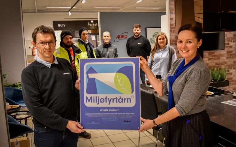 Mann og dame som holder frem et Miljøfyrtårn-diplom med mennesker i bakgrunnen