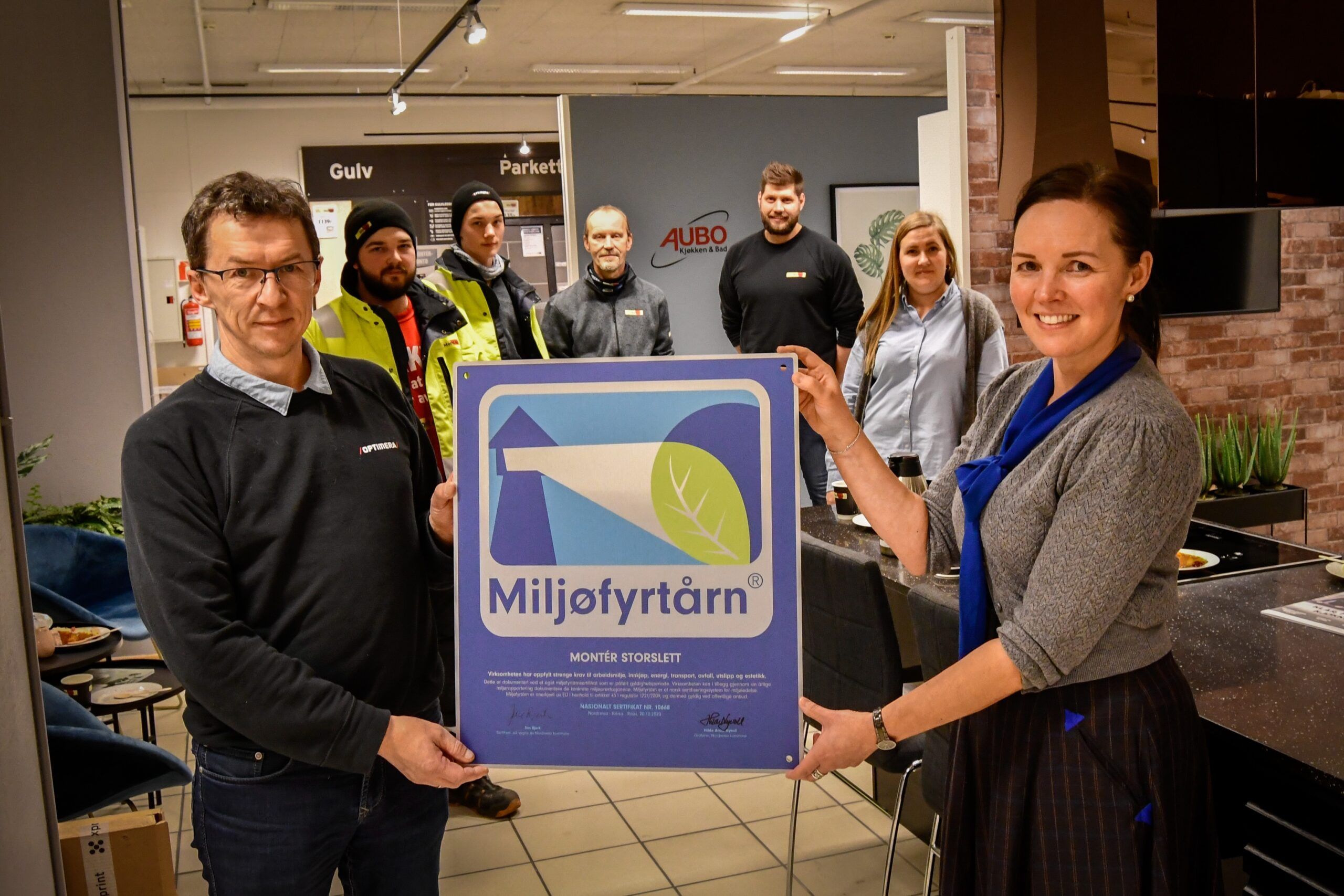 Mann og dame som holder frem et Miljøfyrtårn-diplom med mennesker i bakgrunnen