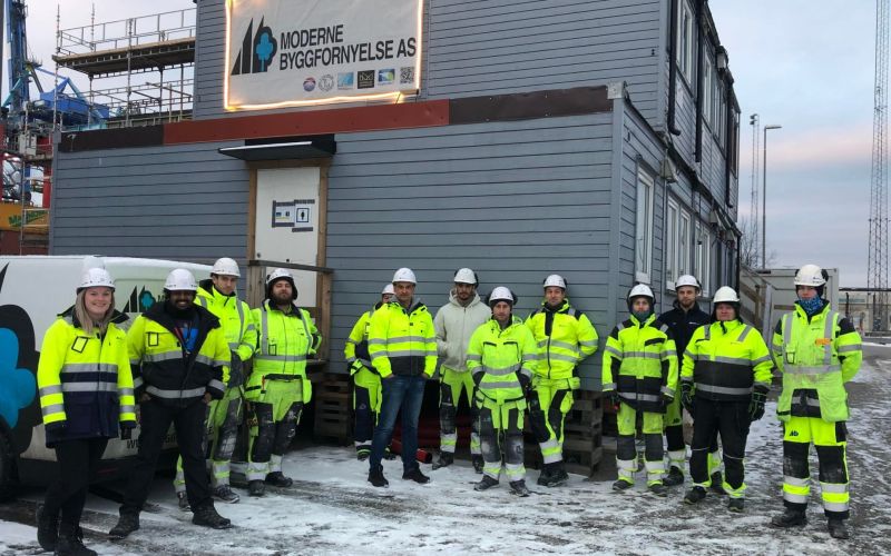 Byggeplass med anleggsarbeidere oppstilt foran varebil og brakker