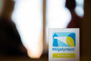 Miljøfyrtårn-plakett i forgrunnen, med menneskeskisser i bakgrunnen.