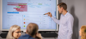 Bilde av mann som peker på tiltaksverktøyet på skjerm foran et møtebord med mennesker