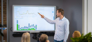 Bilde av mann som peker på grafer på skjerm foran et møtebord med mennesker