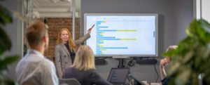 Bilde av dame som peker på grafer på skjerm foran et møtebord med mennesker