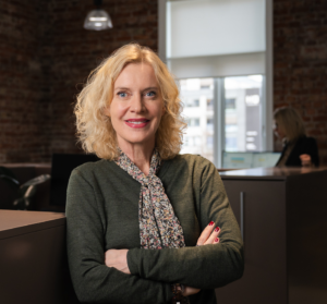 Smilende kvinne i et kontorlandskap. Hun har armene i kors.