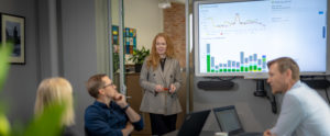 Bilde av dame står ved siden av en skjerm med grafer foran et møtebord med mennesker
