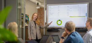 Bilde av dame som peker på grafer på skjerm foran et møtebord med mennesker