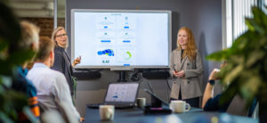 To damer som presenterer på skjerm foran et møtebord med mennesker