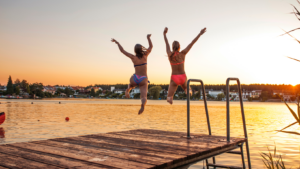 To kvinner som hopper fra en brygge og ut i en sjø. Det er sommer og solnedgang.